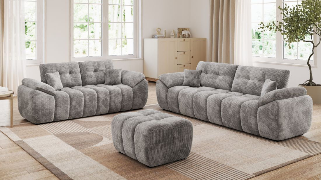 Helles Wohnzimmer mit zwei grauen Sofas und passendem Hocker auf Teppich vor großen Fenstern.
