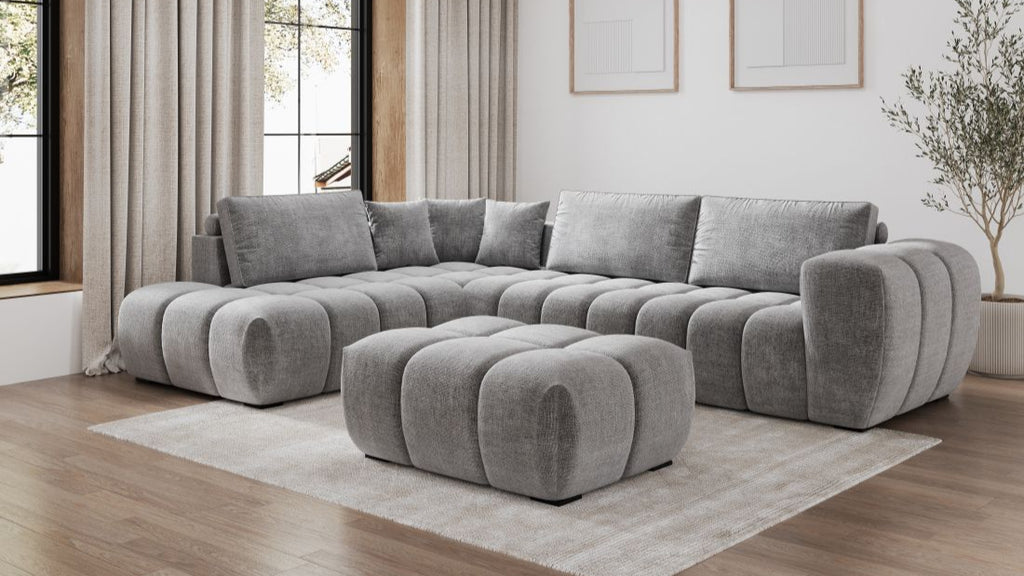 Graues modulares Ecksofa mit passendem Hocker in modernem Wohnzimmer mit großen Fenstern und hellen Vorhängen.